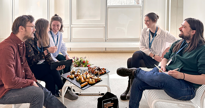 Die Fachgruppe Jungapprobierte sitzt in einem hellen Raum um einen kleinen Tisch mit Snacks und Getränken; sie unterhalten sich in entspannter Atmosphäre